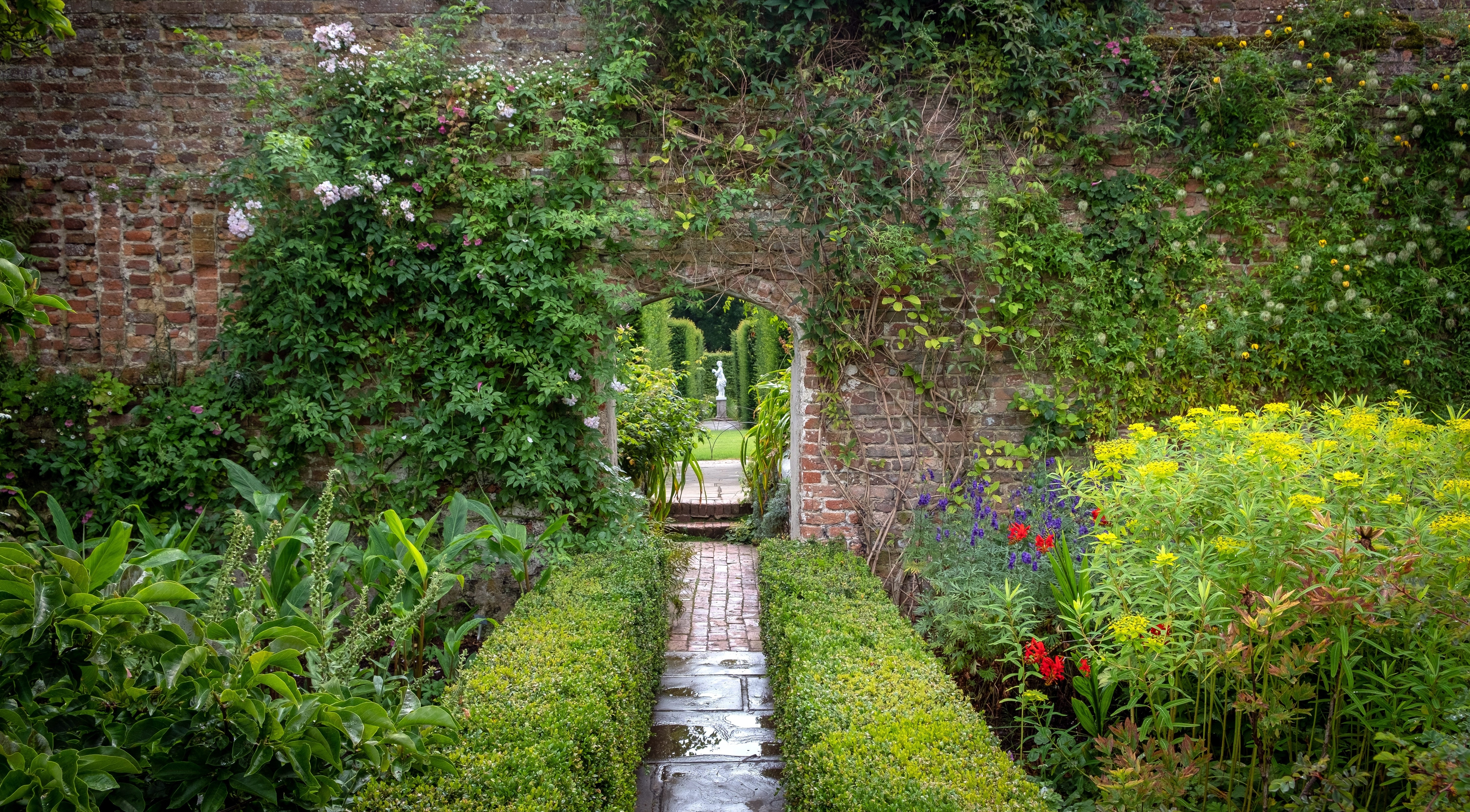 Idyllisk indhegnet have ved Sissinghurst Castle Garden i England med røde og lilla blomster langs en brolagt sti og en historisk muråbning