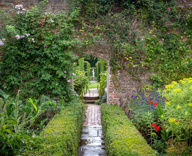 Idyllisk indhegnet have ved Sissinghurst Castle Garden i England med røde og lilla blomster langs en brolagt sti og en historisk muråbning