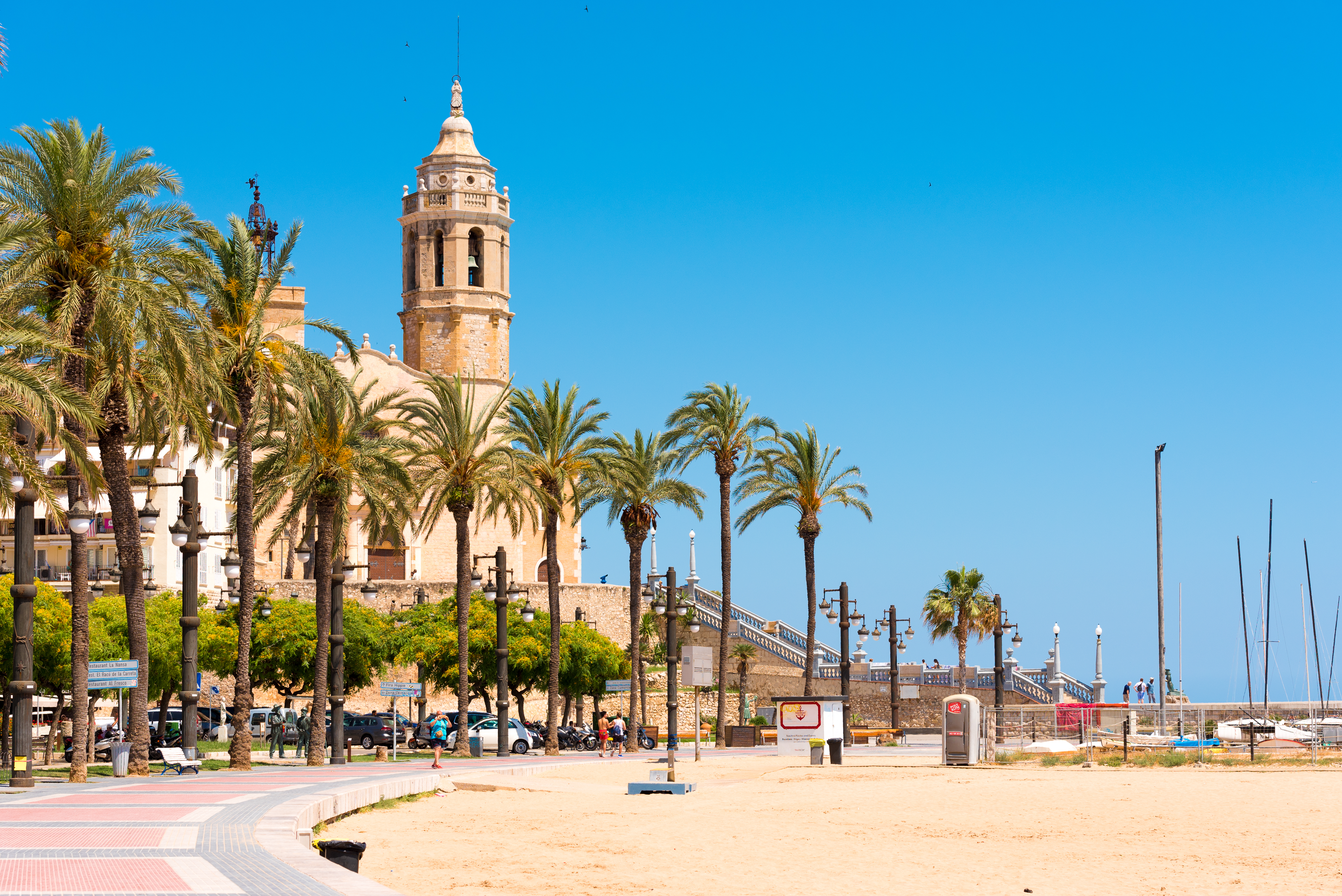 Populært feriedestination Sitges med sandstrand, palmelinede promenade og den smukke Sant Bartomeu kirke under klar blå himmel, Spanien