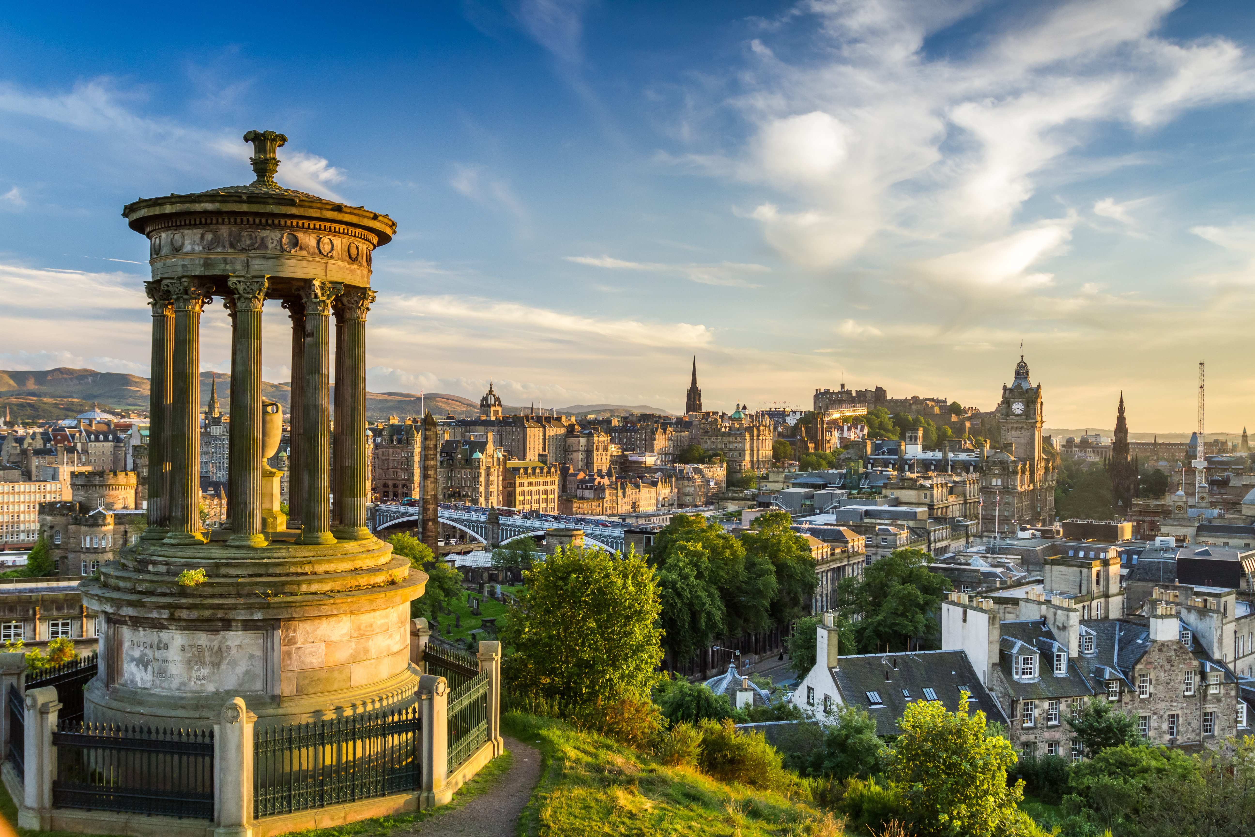 Panoramaudsigt over Edinburgh med klassisk monument på Calton Hill og Edinburgh Castle i horisonten ved solnedgang