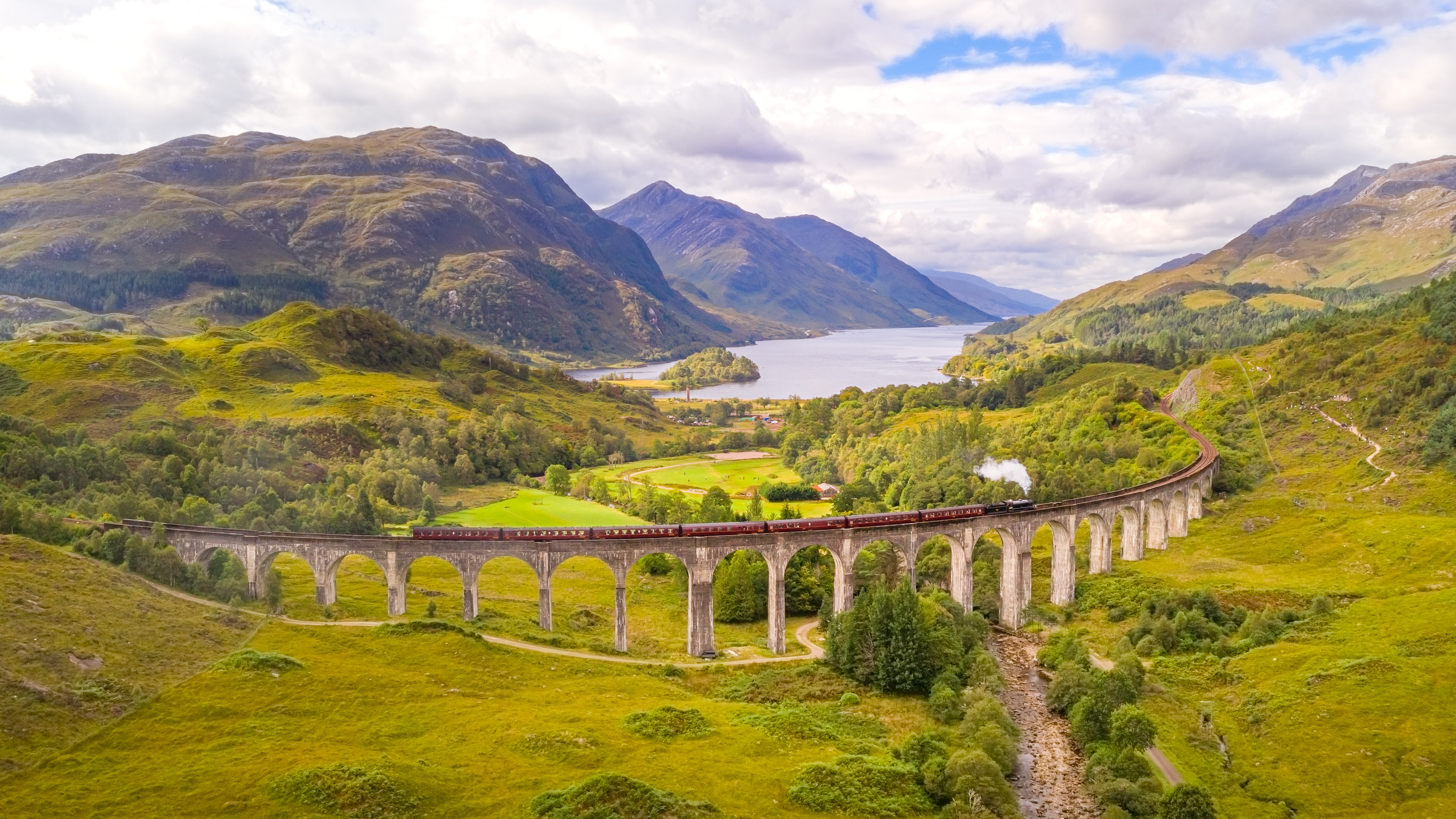 Det berømte Glenfinnan Viadukt i Skotland set fra luften med et damptog der krydser broen omgivet af grønne bjerge og Loch Shiel