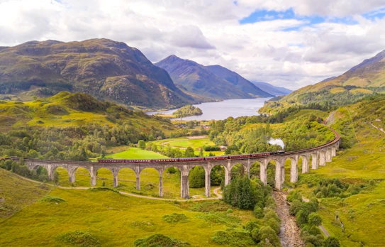 Det berømte Glenfinnan Viadukt i Skotland set fra luften med et damptog der krydser broen omgivet af grønne bjerge og Loch Shiel