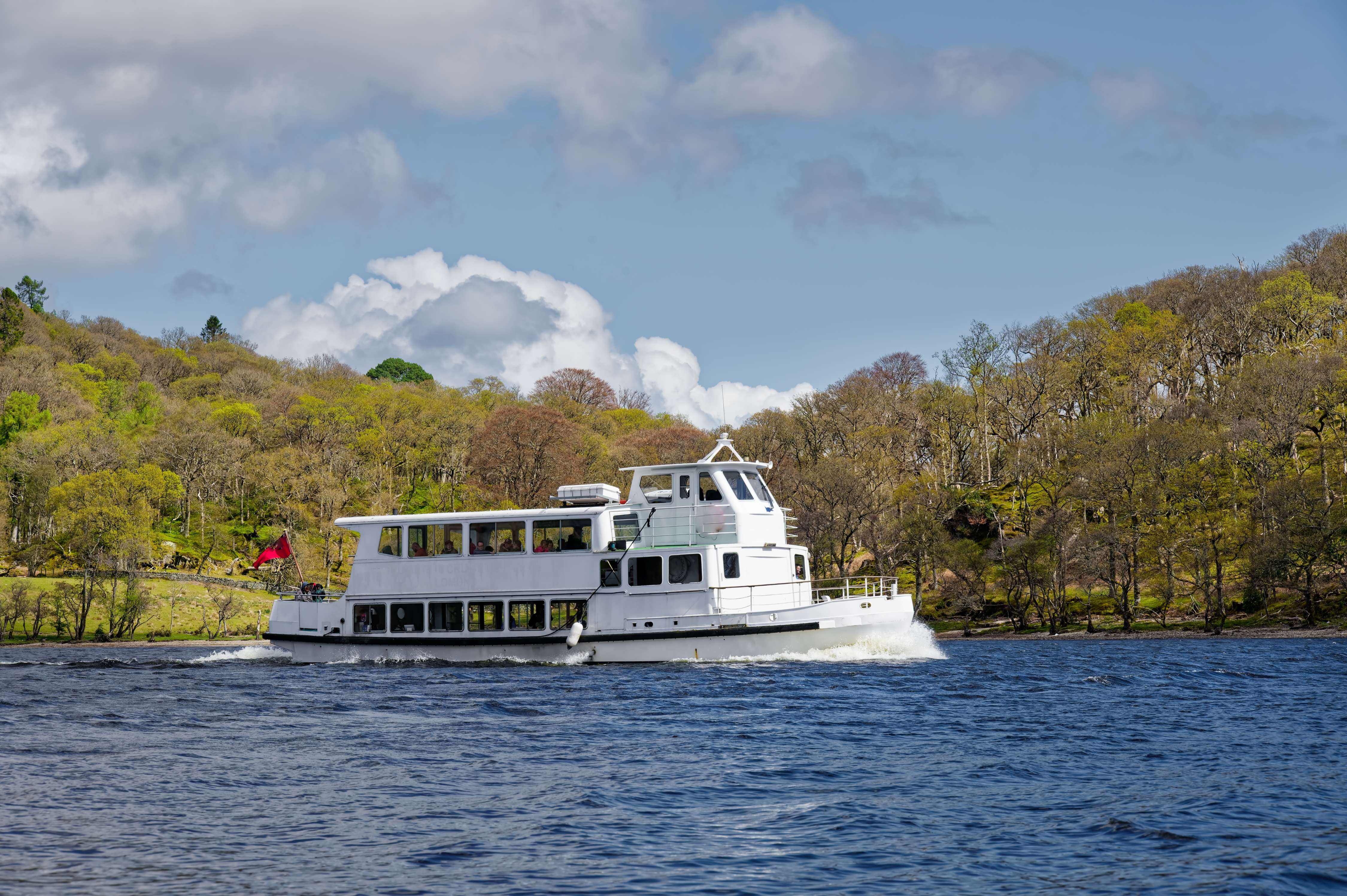 Hvid turistbåd på den smukke Loch Lomond i Skotland omgivet af frodig skov og det karakteristiske skotske højlandslandskab