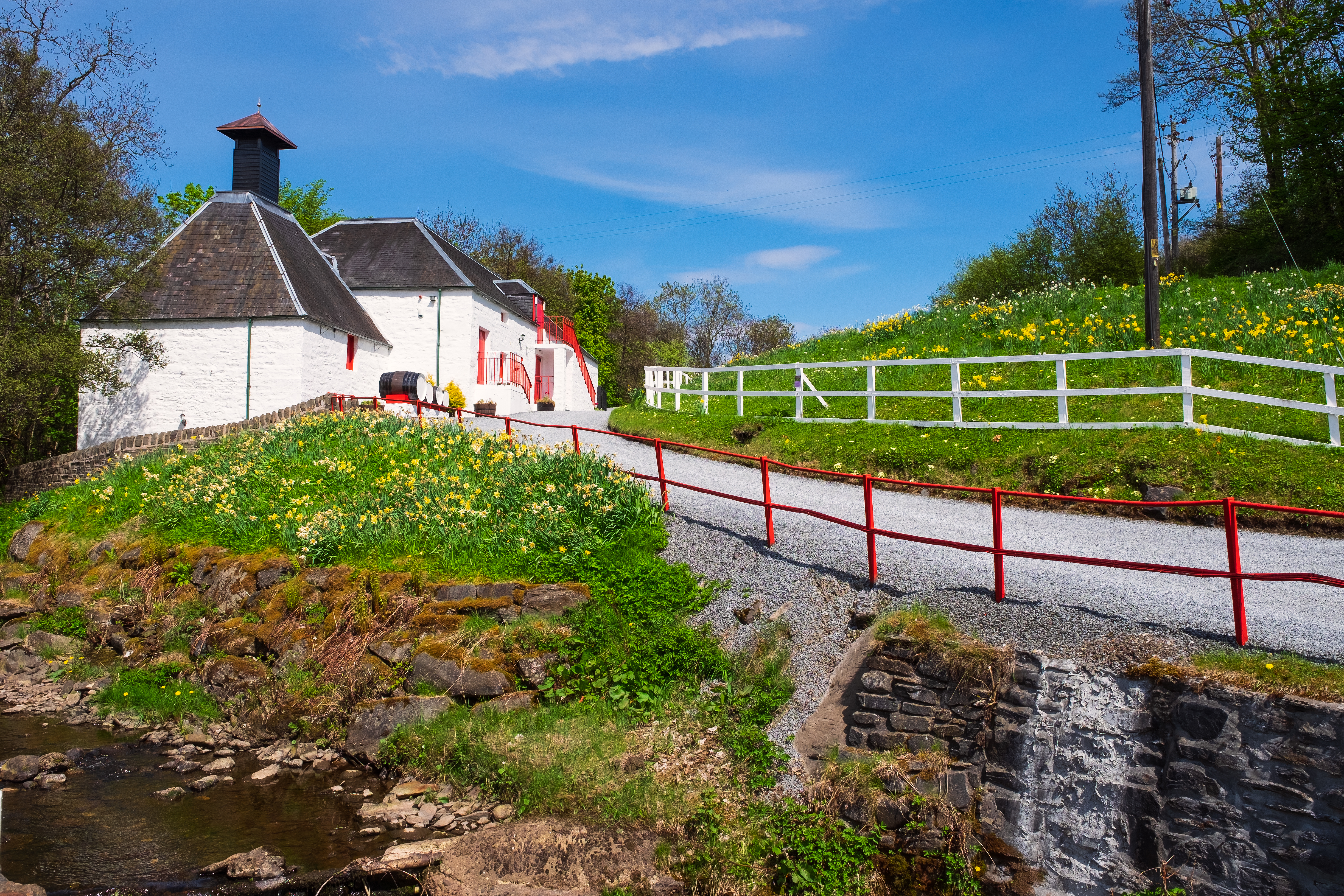Smukt traditionelt destilleri i de skotske Highlands med hvide mure, røde detaljer og pagodetag omgivet af frodig natur og vilde blomster langs en lille å nær Pitlochry