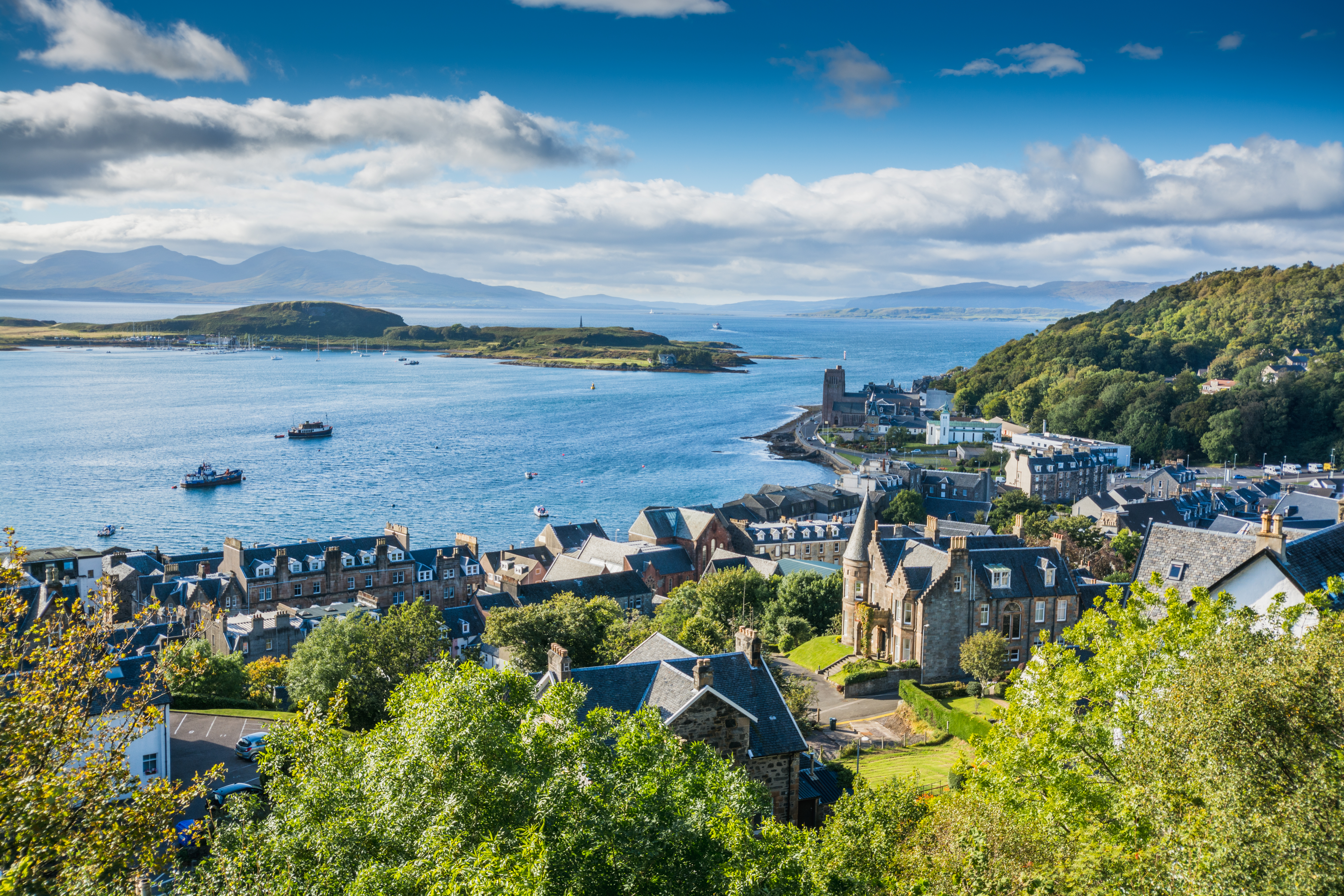 Spektakulært luftbillede af den maleriske kystby Oban i Skotland, med dens charmerende havn, historiske bygninger og kirke, omgivet af spredte træer og det blå hav, der strækker sig mod horisonten