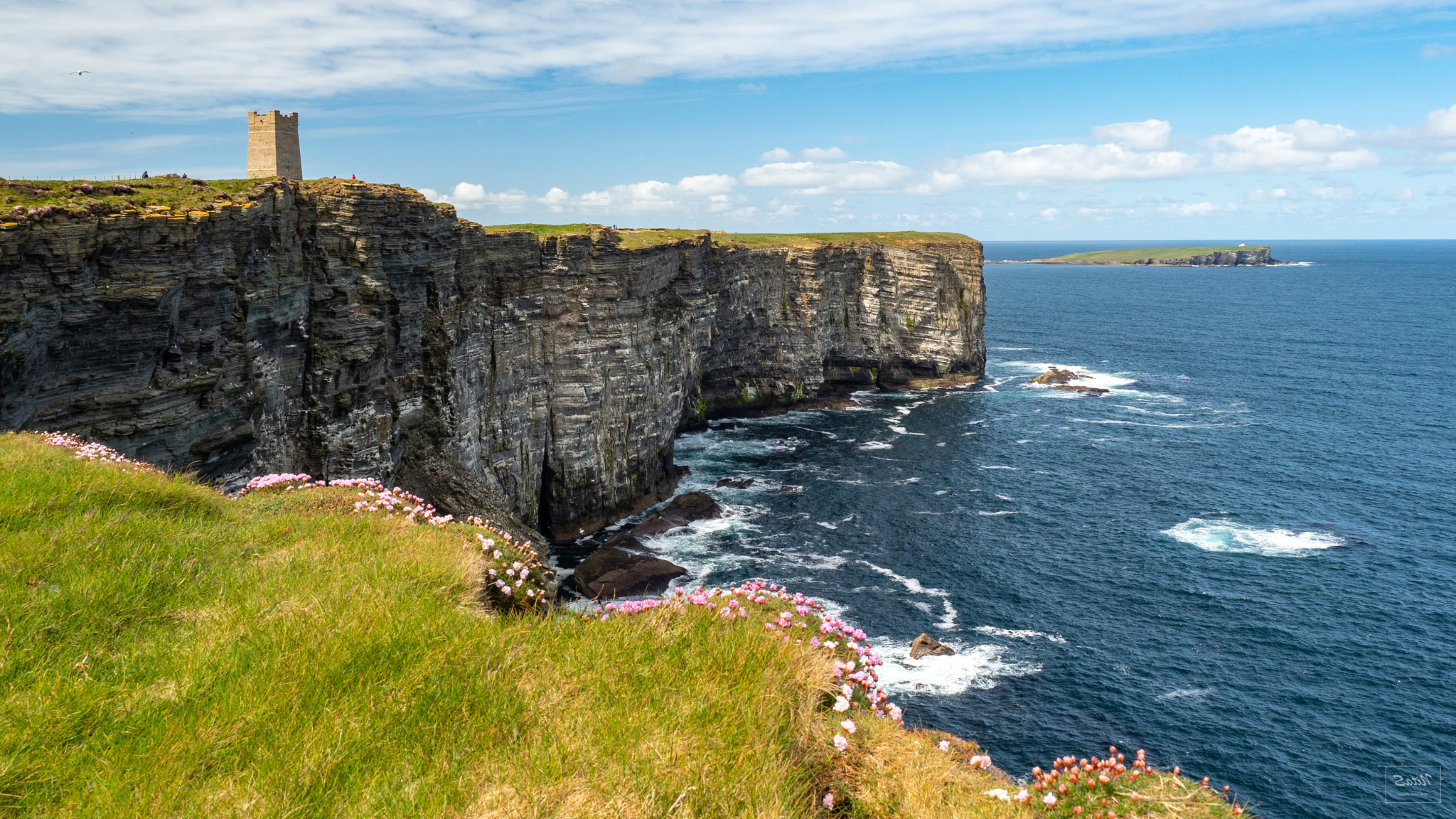 Storslåede klipper og det blå hav ved den skotske nordkyst med ruiner af gammelt tårn og den ubeboede ø Stroma i Orkney-øgruppen i baggrunden