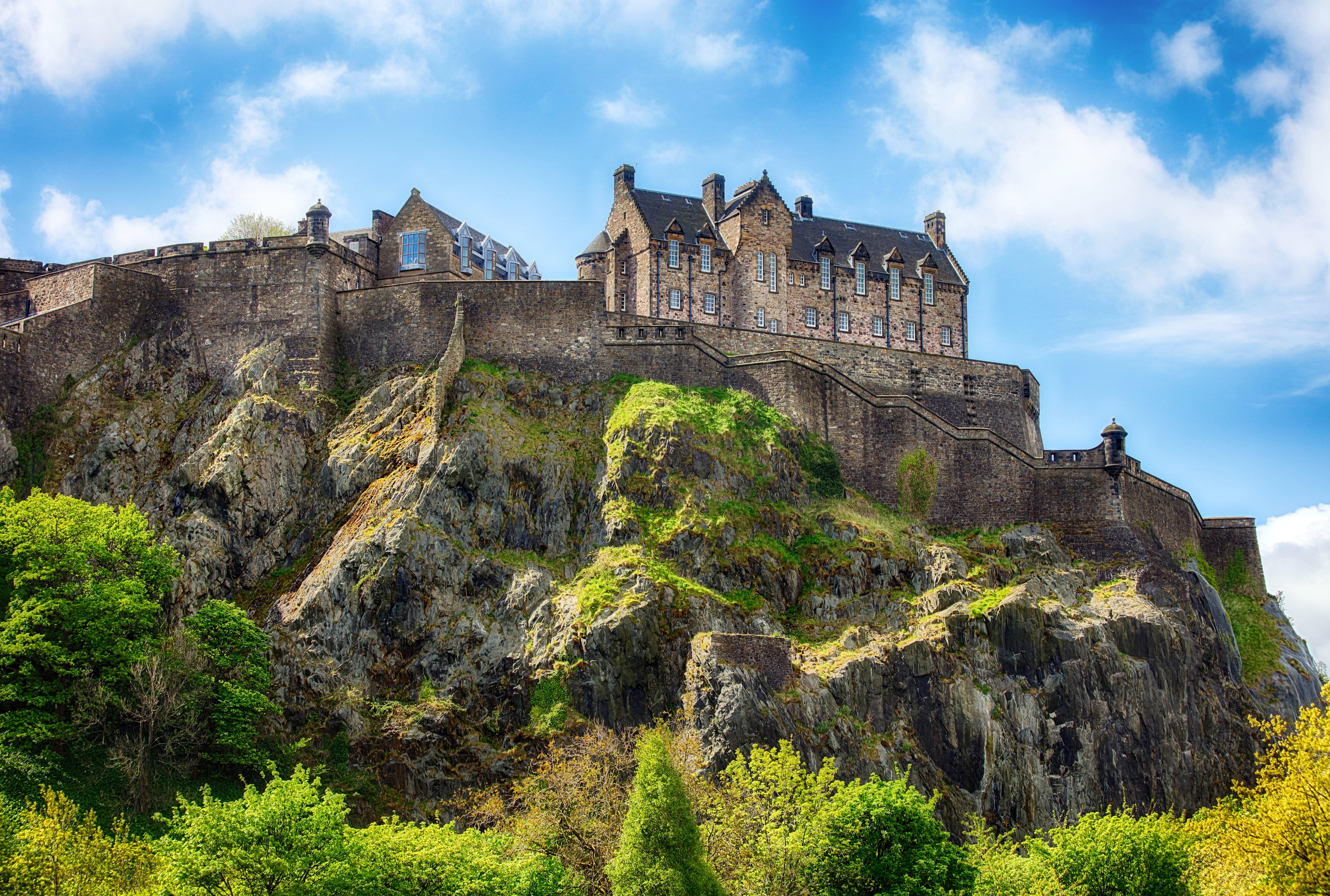 Det storslåede Edinburgh Castle på toppen af Castle Hill med sine karakteristiske stenbyggede fæstningsmure, omgivet af grønne bakker under en klar blå skotsk himmel
