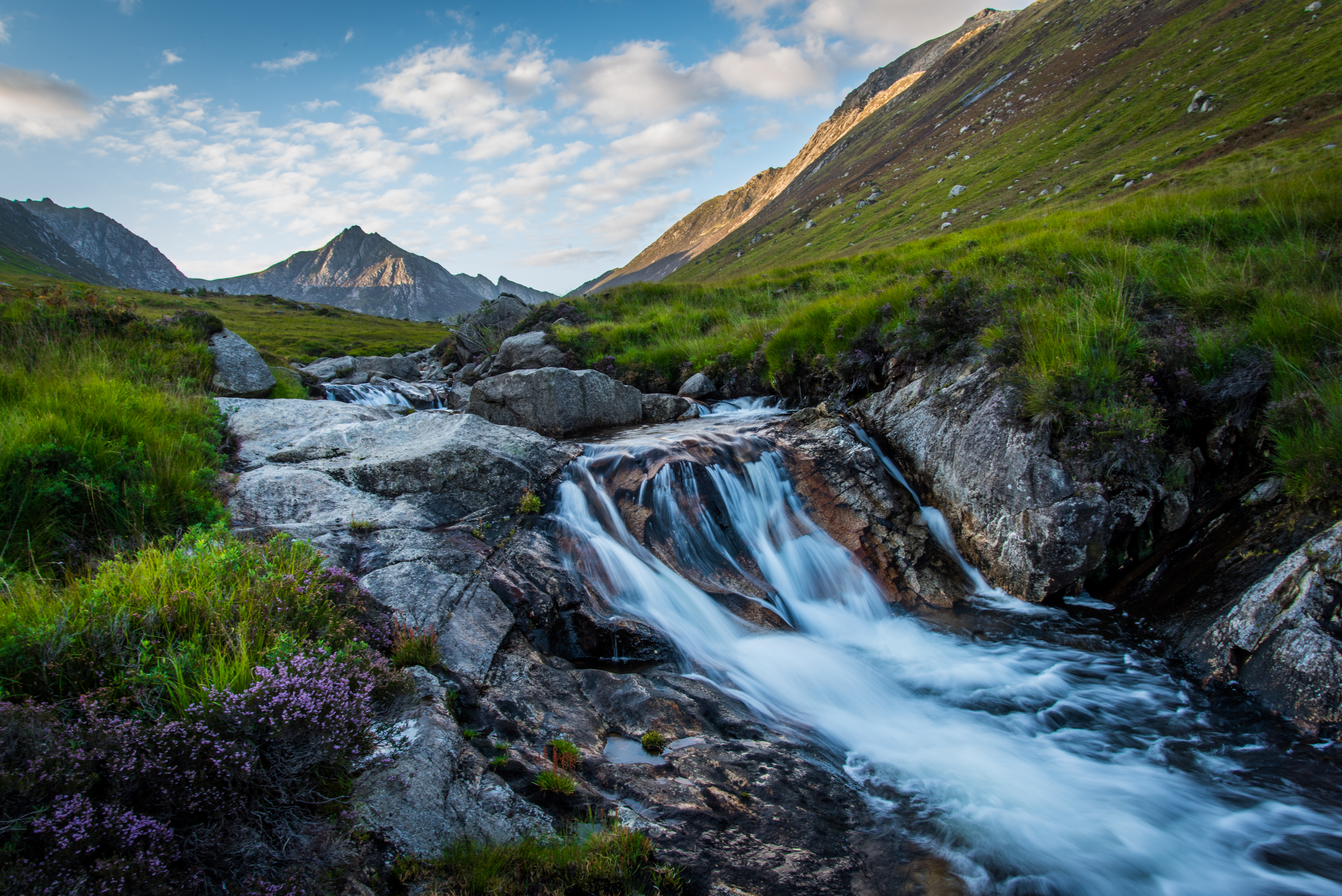 Vild skotsk natur med et smukt vandfald over klipper, lilla hedeblomster og grønne bjergdale i Highlands under en blå himmel