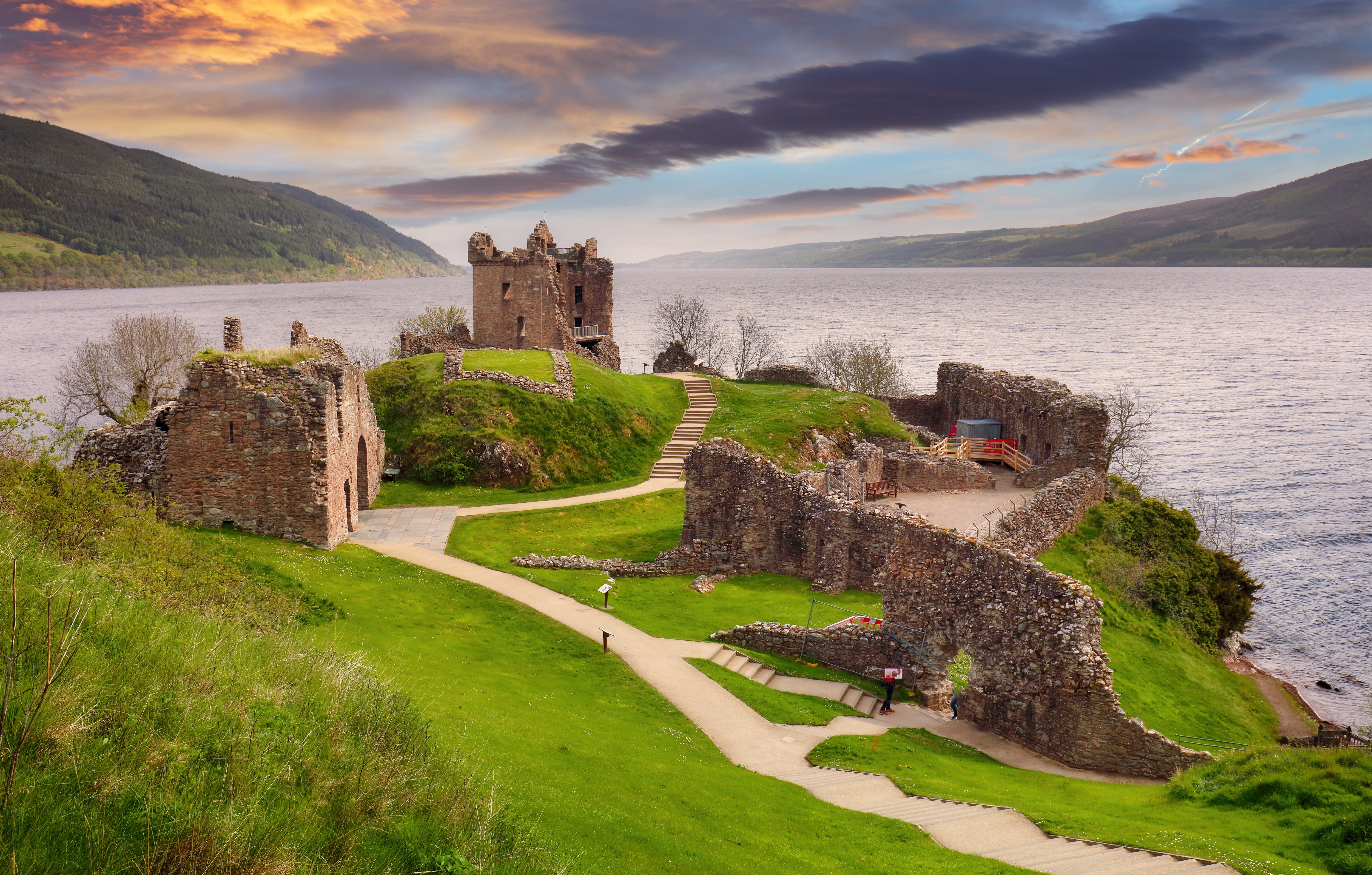 Den historiske fæstning Urquhart Castle med den ikoniske Loch Ness i baggrunden under en betagende rød solnedgang i Skotland