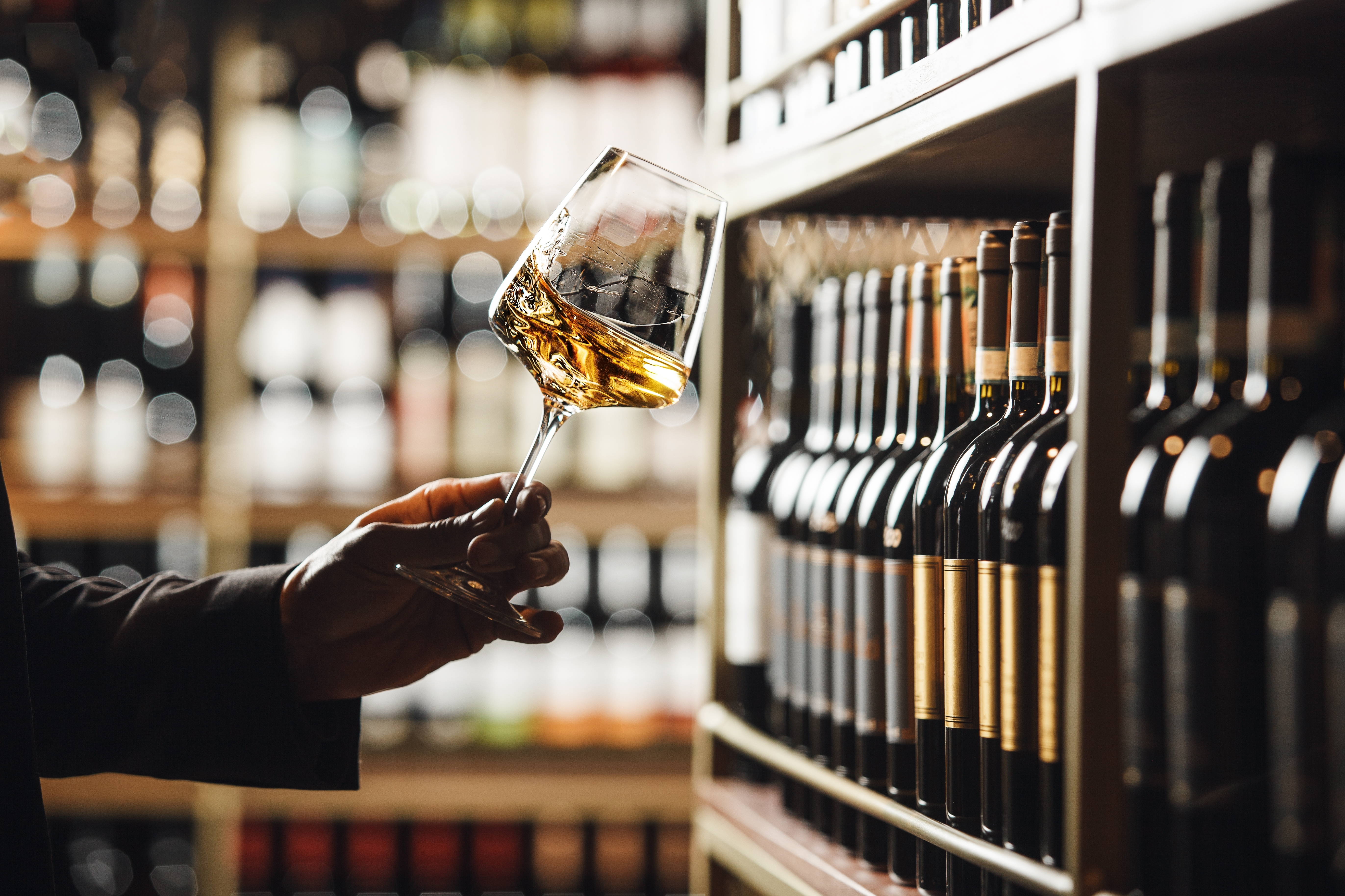 Nærbillede af sommelier der swirler og smagsanalyser hvidvin omgivet af vinflasker i vinkælder
