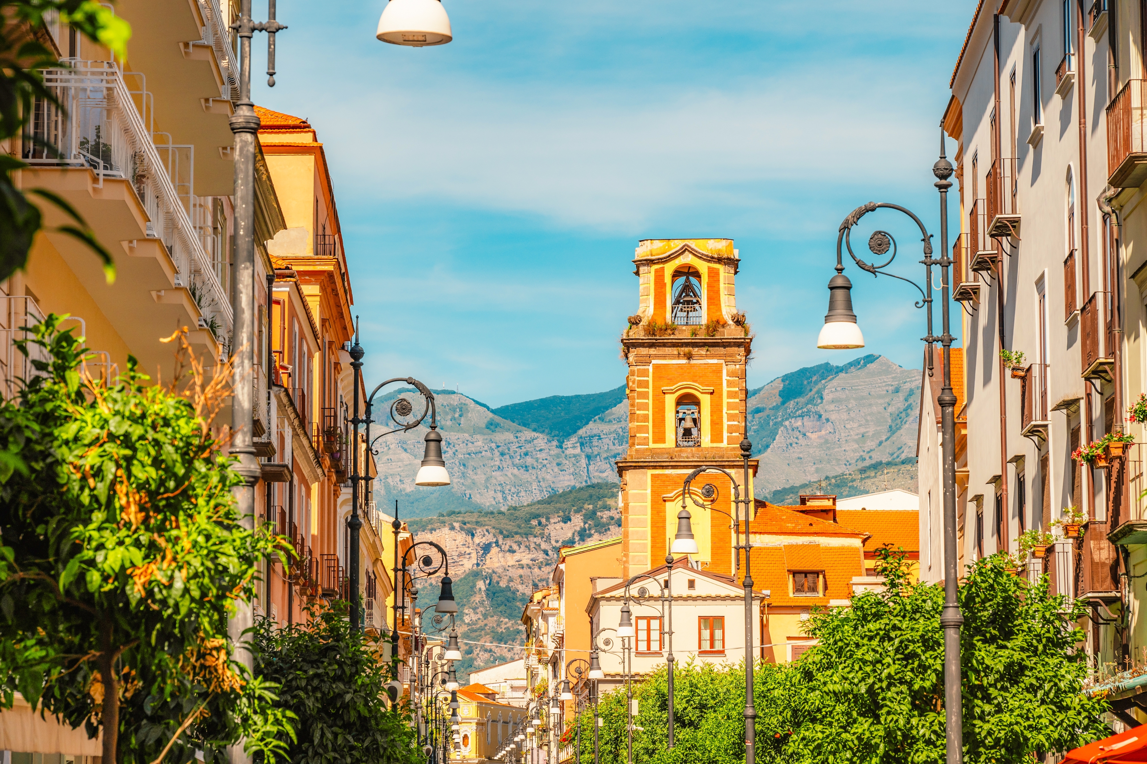 Stemningsfuld gade i Sorrento med gylden kirketårn, pastelfarvede bygninger og majestætiske bjerge i baggrunden under azurblå himmel
