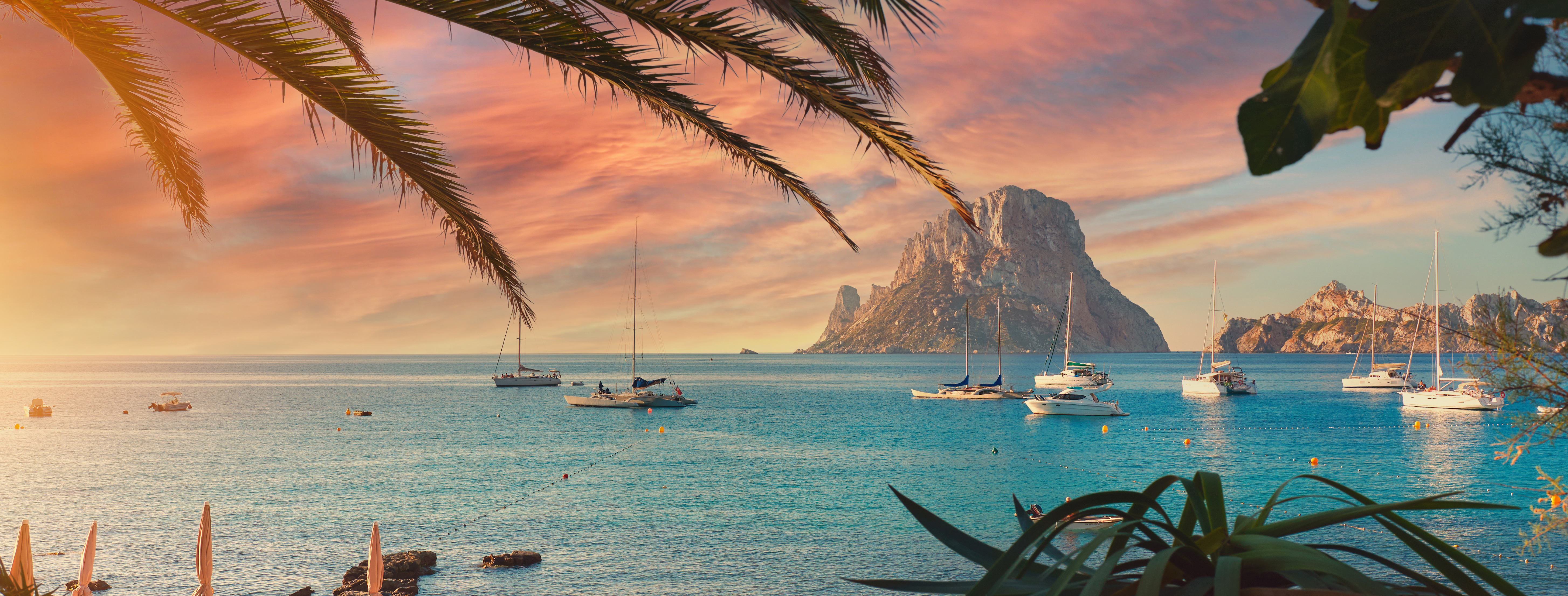 Malerisk spansk kyst med sejlbåde og dramatisk Es Vedra klippeformation ved solnedgang, Ibiza