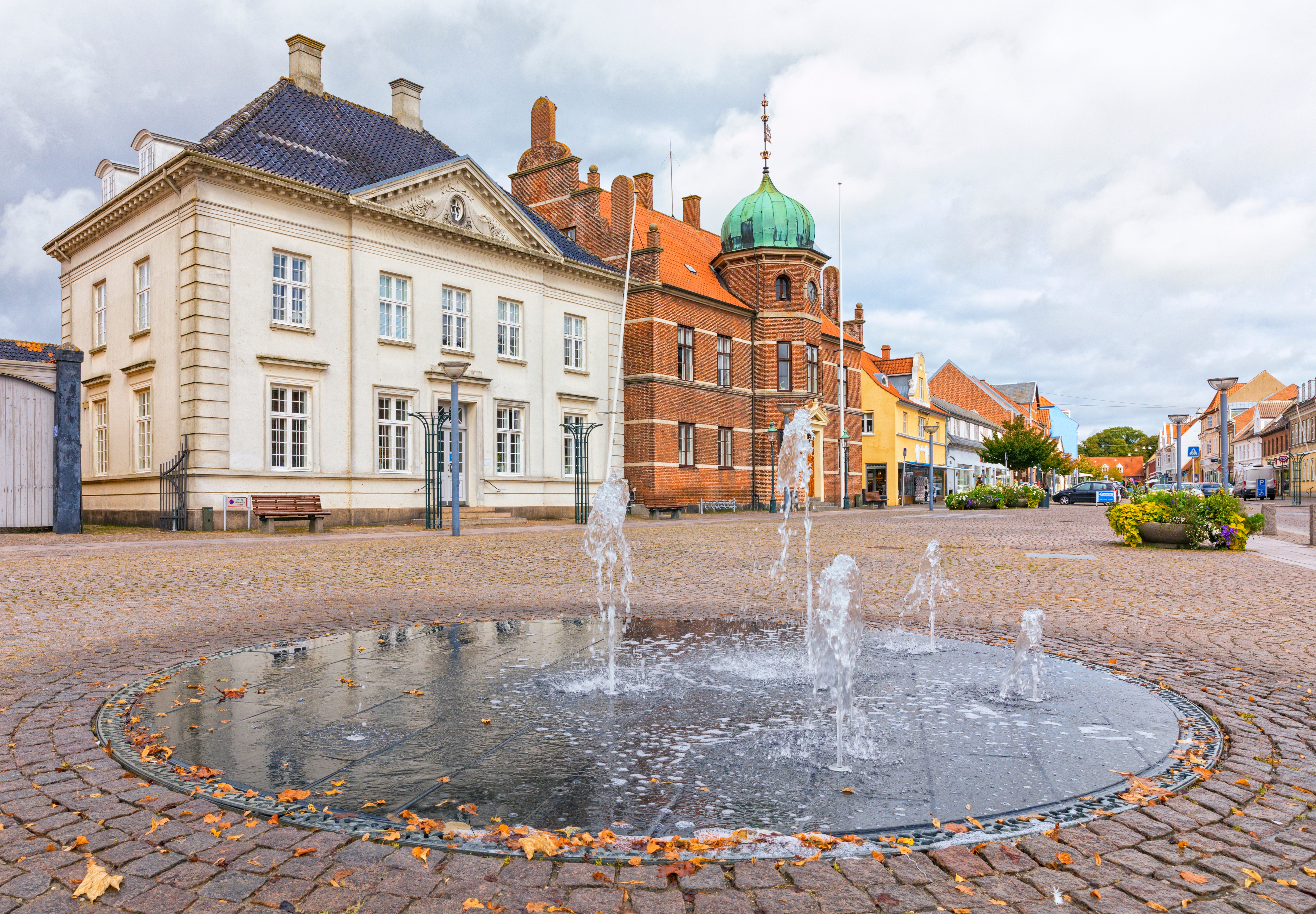 Hyggelig torveplads i Stege på Møn med springvand, historisk sparekasse og rødmuret rådhus under en overskyet himmel