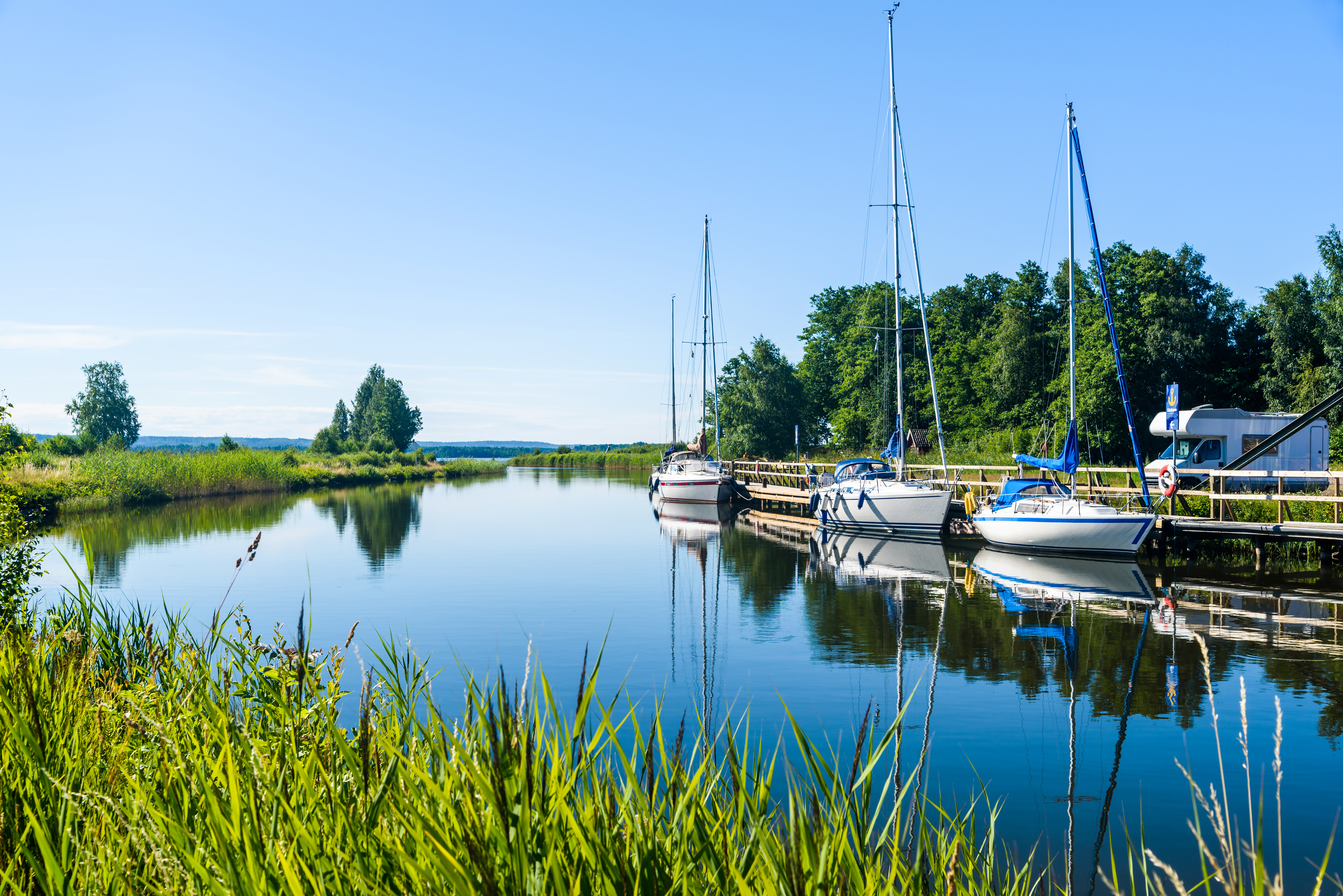 Rolig svensk kanal med fortøjede sejlbåde og træbro i smukt grønt sommerlandskab