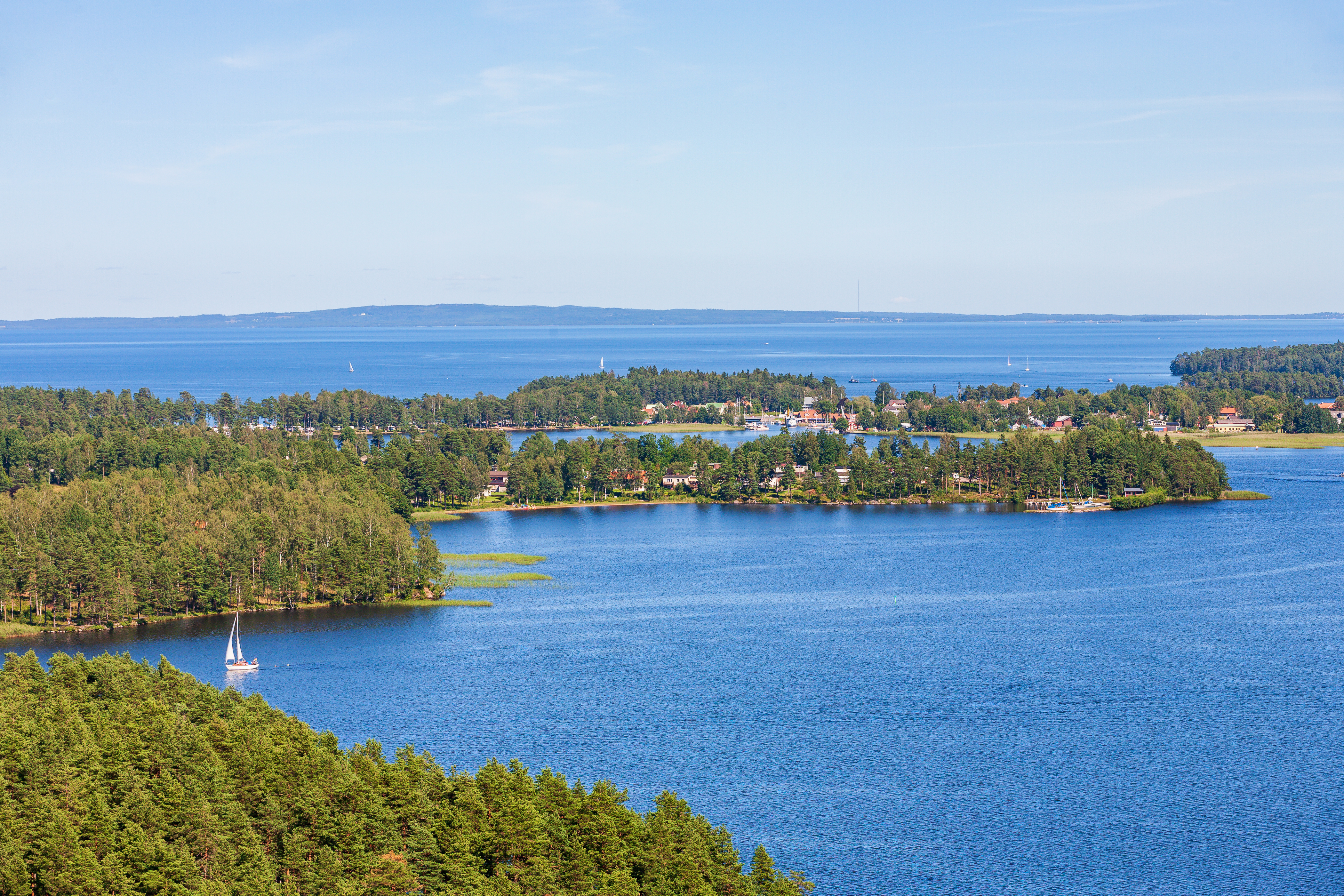 Naturskønt luftfoto af en hvid sejlbåd på Vättern-søen ved Karlsborg i Sverige omgivet af grønne fyrreskovsbeklædte øer og klar blå sommerhimmel