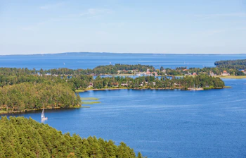 Naturskønt luftfoto af en hvid sejlbåd på Vättern-søen ved Karlsborg i Sverige omgivet af grønne fyrreskovsbeklædte øer og klar blå sommerhimmel
