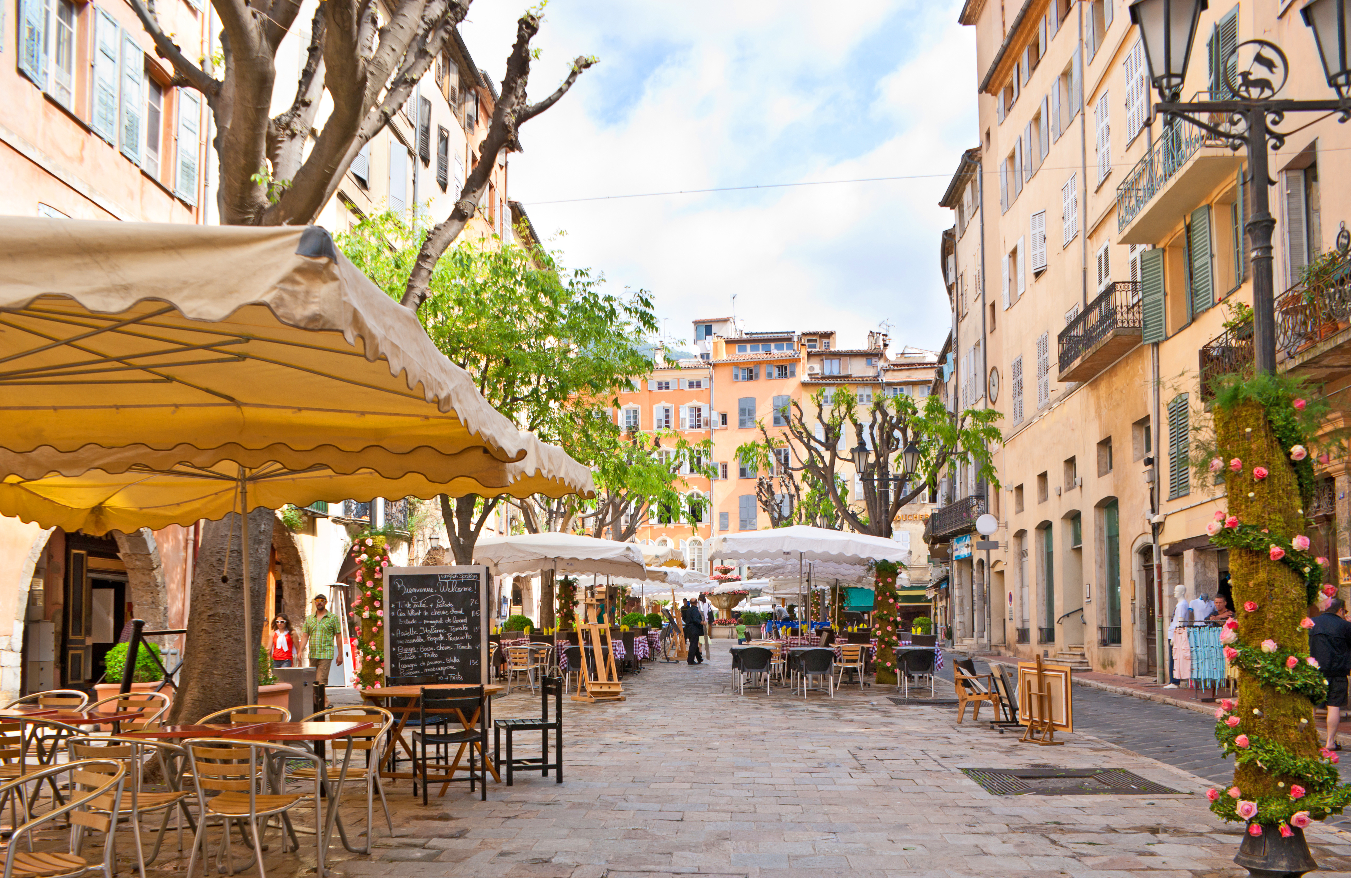 Malerisk café-torv i den historiske by Grasse i Sydfrankrig med gule parasols, stenbygninger og blomstrende roser i det solrige Provence