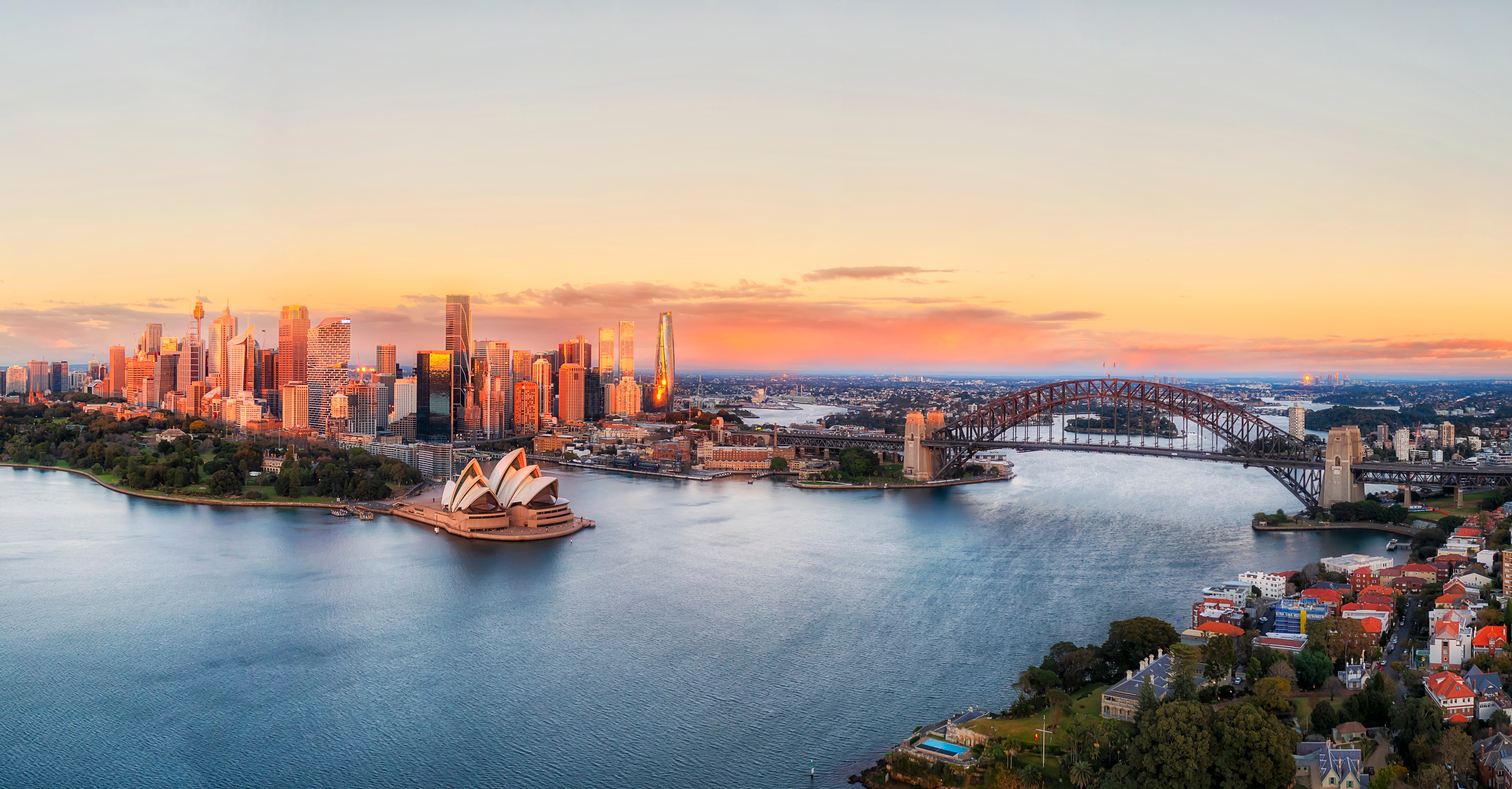 Imponerende panoramabillede af Sydney ved solopgang, hvor det gyldne lys fremhæver Operahusets sejl, Harbour Bridge og byens moderne skyline mod den blå havnefront