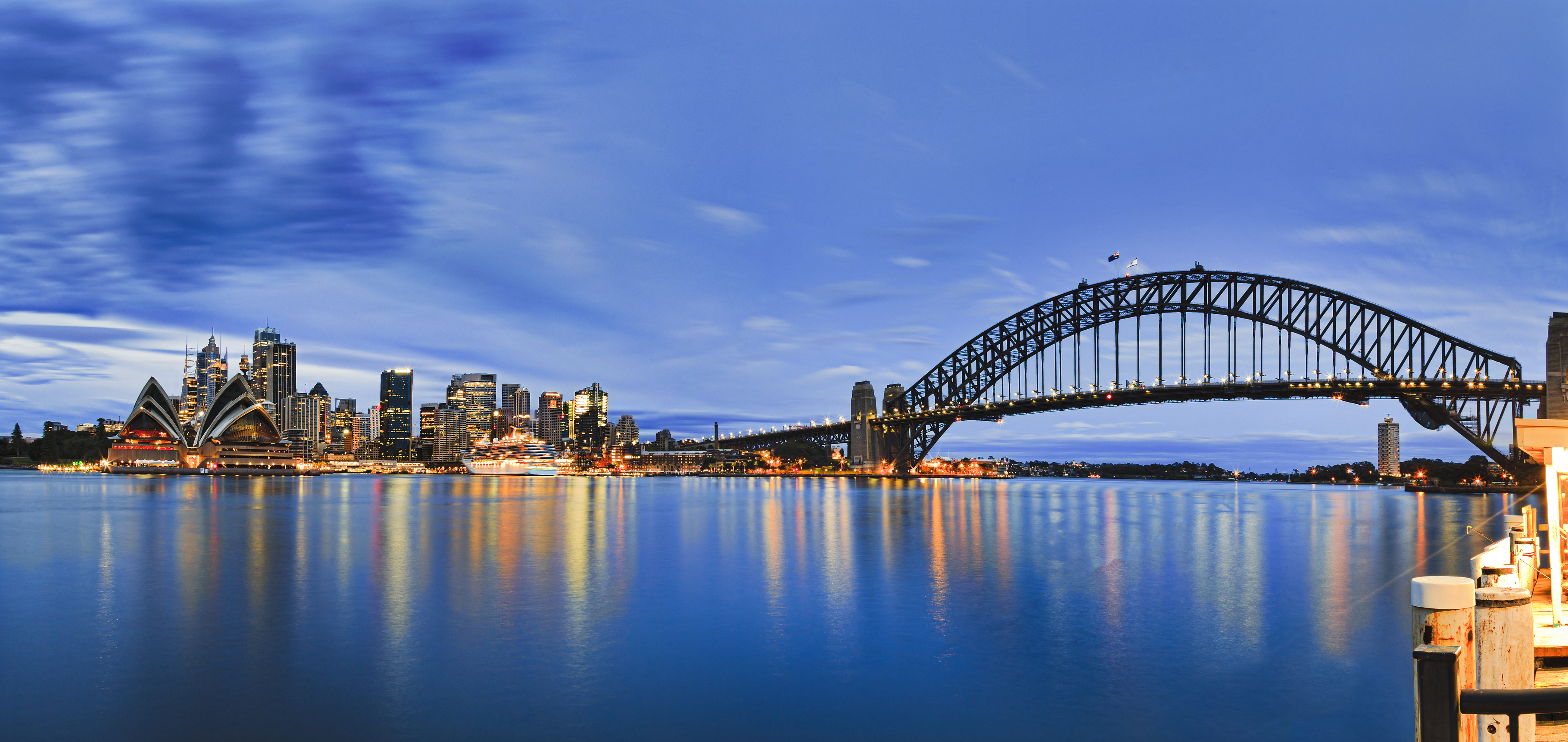 Fascinerende morgenvy over Sydney med den karakteristiske Harbour Bridge og CBD-skyskrabere badet i blåt solopgangslys. Operahuset og passagerterminalen ved Circular Quay spejler sig i havnens vand.