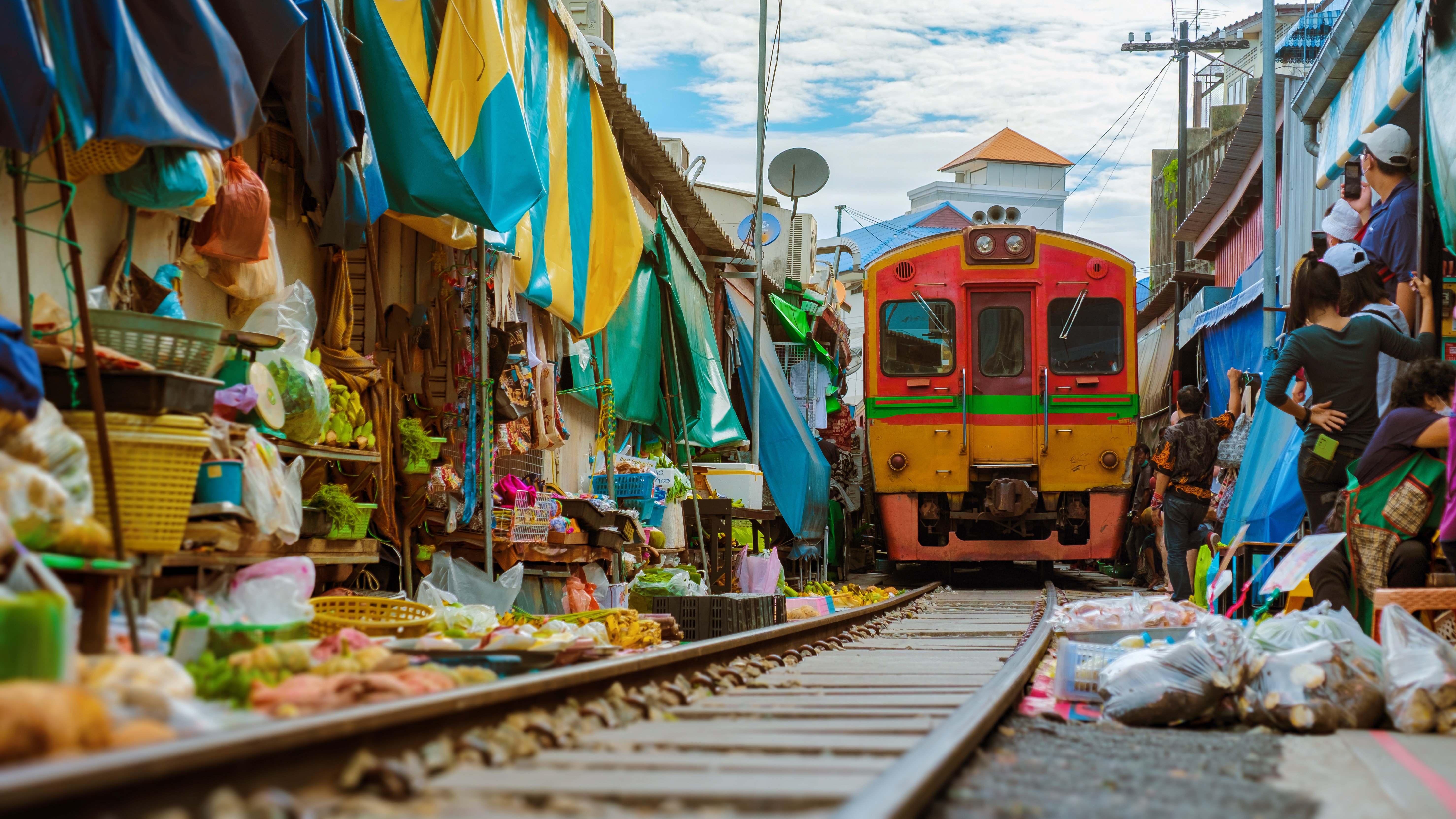 Spændende togmarked i Mae Klong, Thailand, hvor et farverigt lokomotiv kører tæt forbi sælgernes boder med friske varer langs jernbanesporene