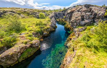 Turkisblåt krystalklart vand strømmer gennem den smukke Silfra-kløft i Thingvellir Nationalpark på Island, omgivet af dramatiske klippeformationer og grøn sommernatur