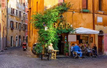 Romantisk smal gade i Trastevere-distriktet i Rom med farverige bygninger, en intim udendørs café og frodige planter, der indfanger den særlige romerske stemning i et af byens mest karakteristiske kvarterer