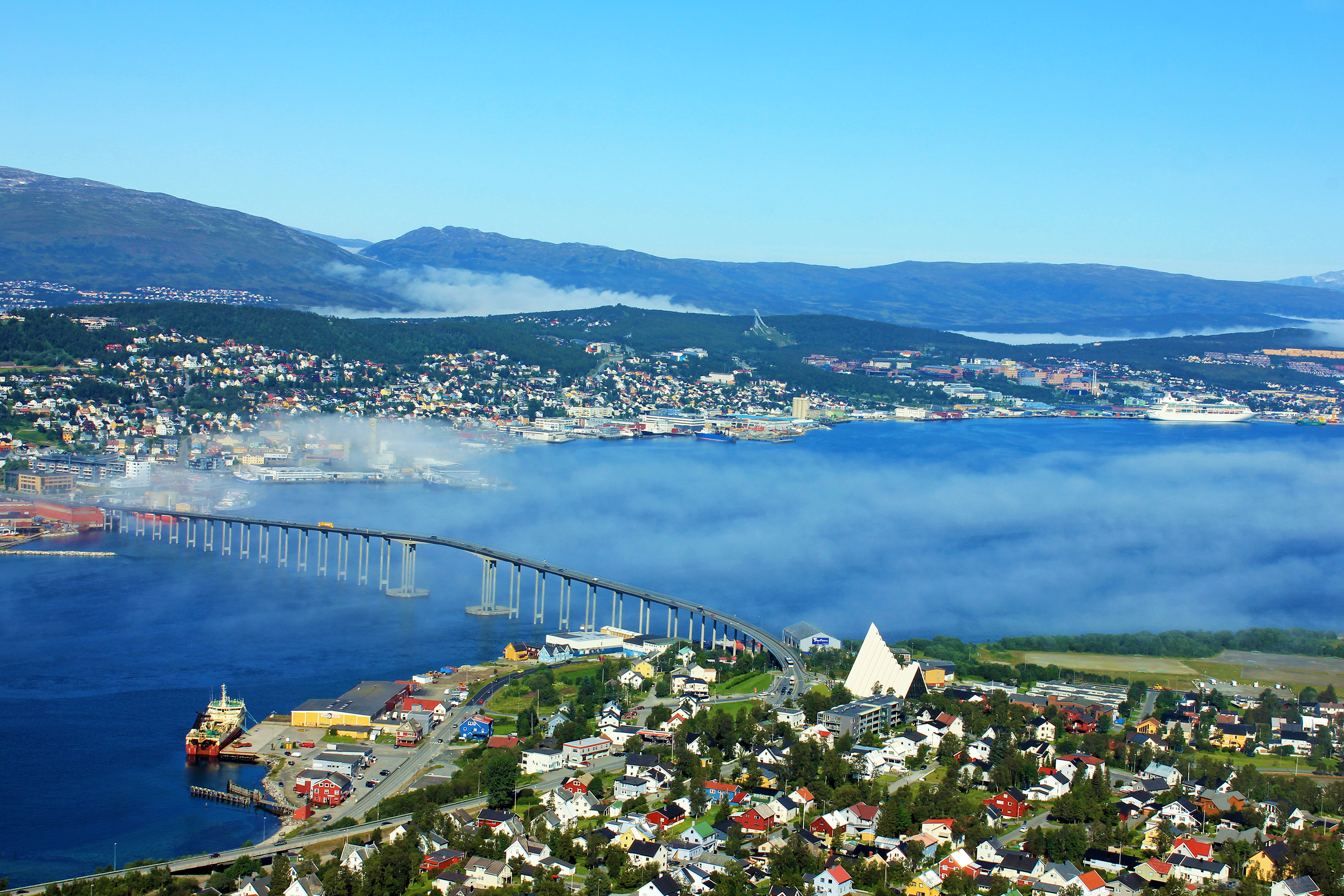 Smukt luftfoto af Tromsø i Nordnorge med den lange bro over fjorden, havnen med skibe og de omkringliggende fjelde dækket af let tåge på en klar sommerdag