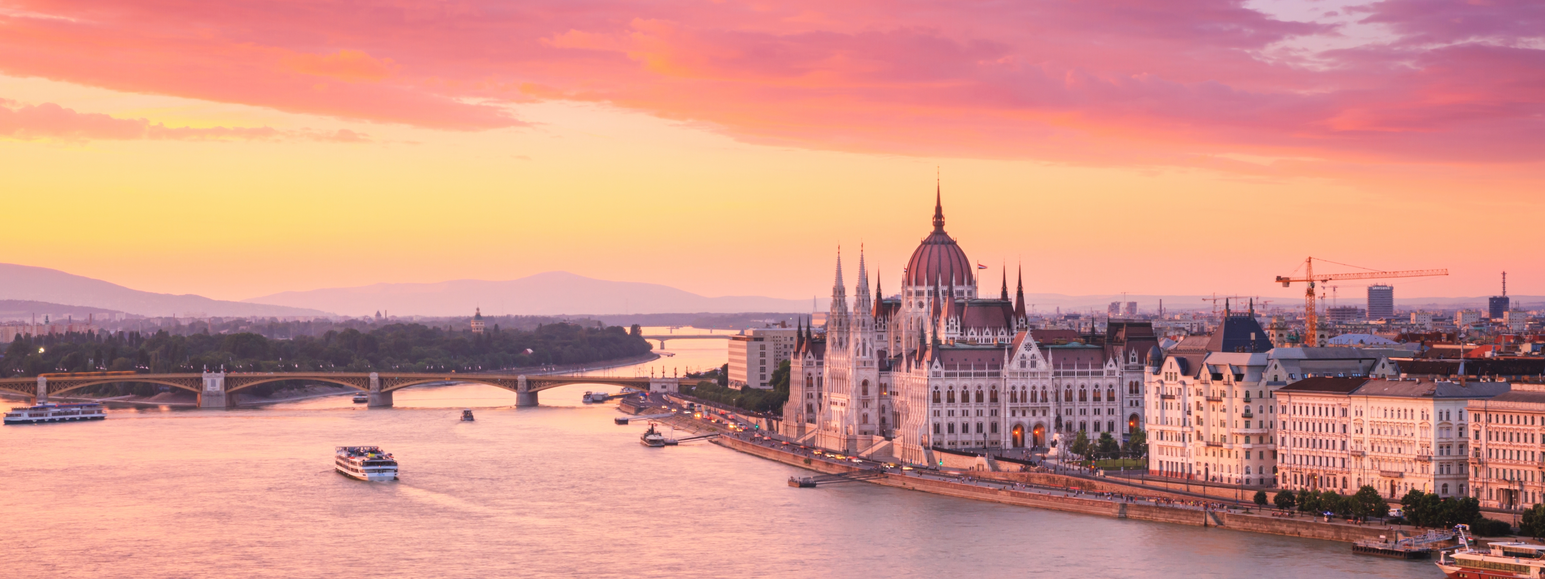 Imponerende solnedgangsudsigt over Budapests historiske centrum med den gotiske parlamentsbygning, den blå Donauflod og den ikoniske Margaretbro i Ungarn