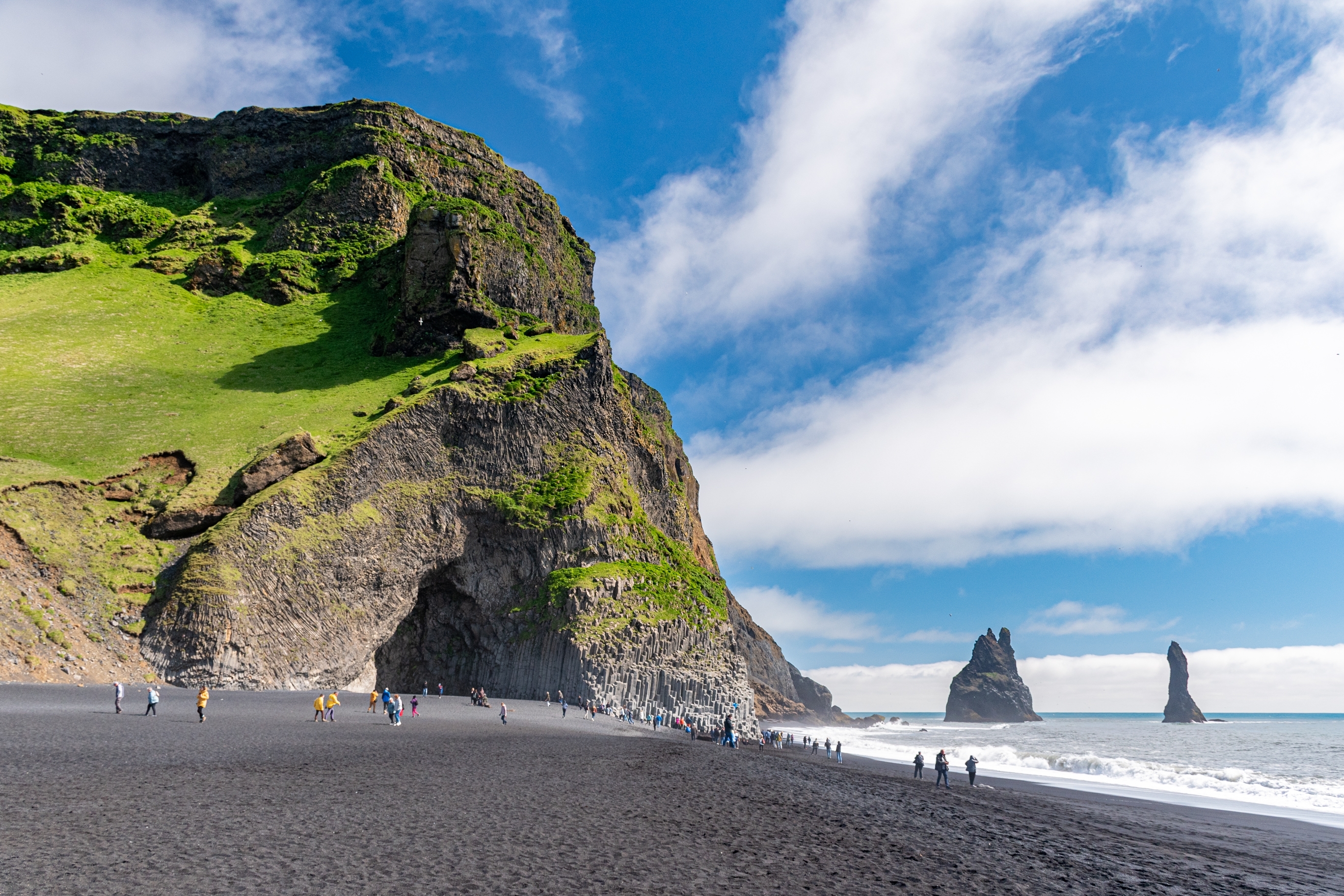 Reynisfjara strand ved Vik på Island med sort sand, grønne klipper og Reynisdrangar søjler rejser sig fra havet under en delvist skyet blå himmel
