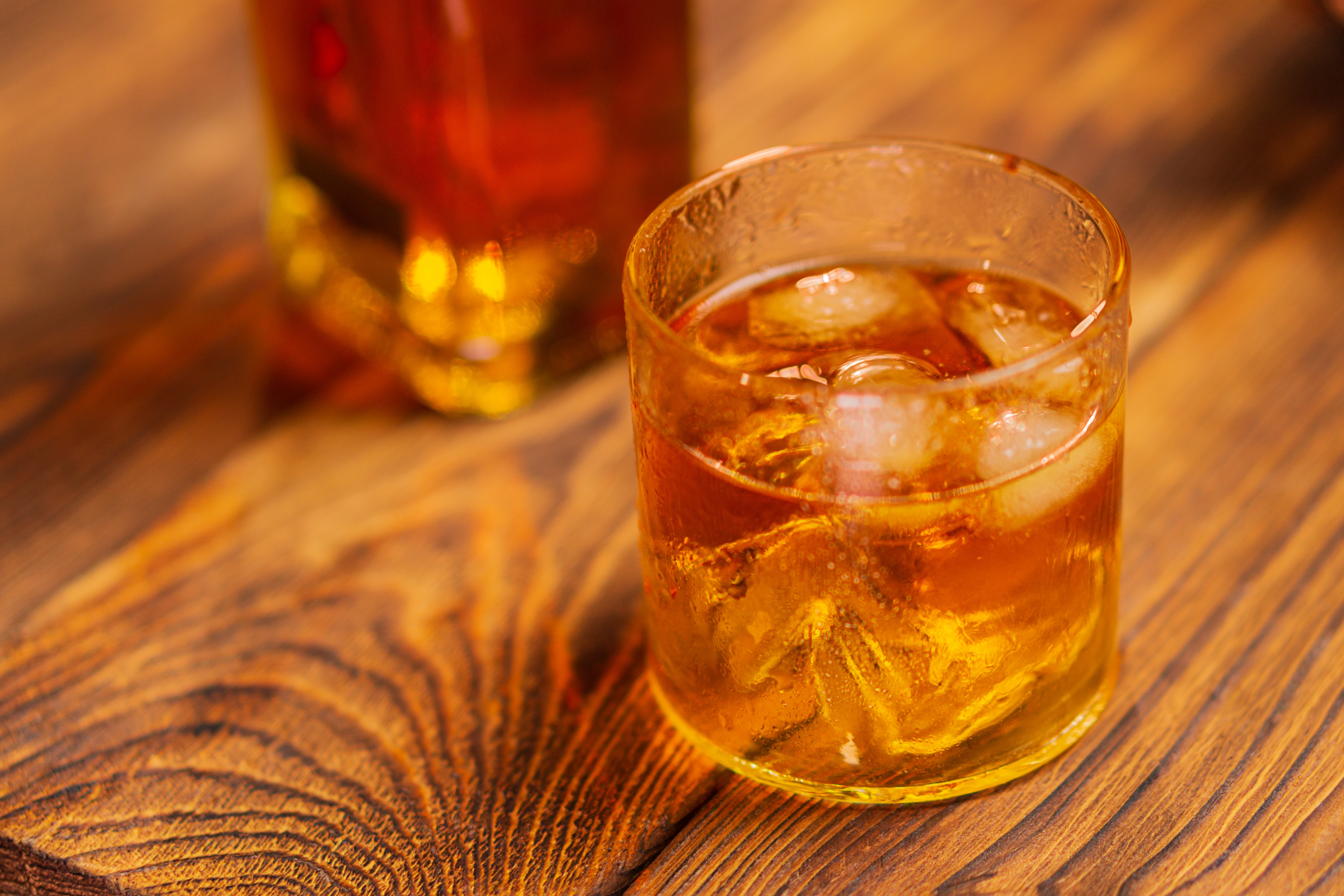 Gyldent whisky-glas med isterninger på et rustikt pinjetre-bord med sløret flaske i baggrunden
