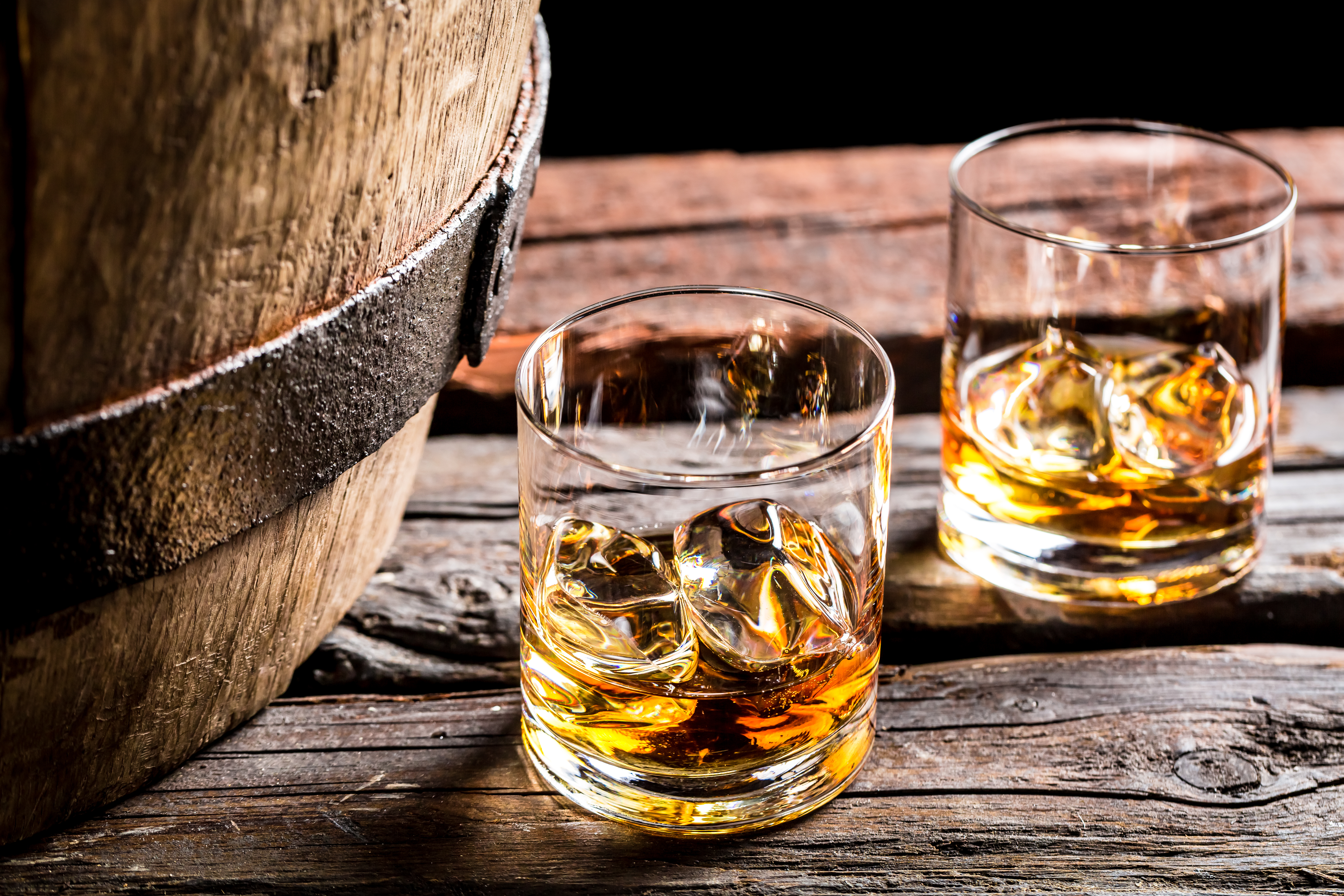 To glas med gylden whisky og isklumper på et rustikt træbord ved siden af et gammelt træfad i en traditionel kælder