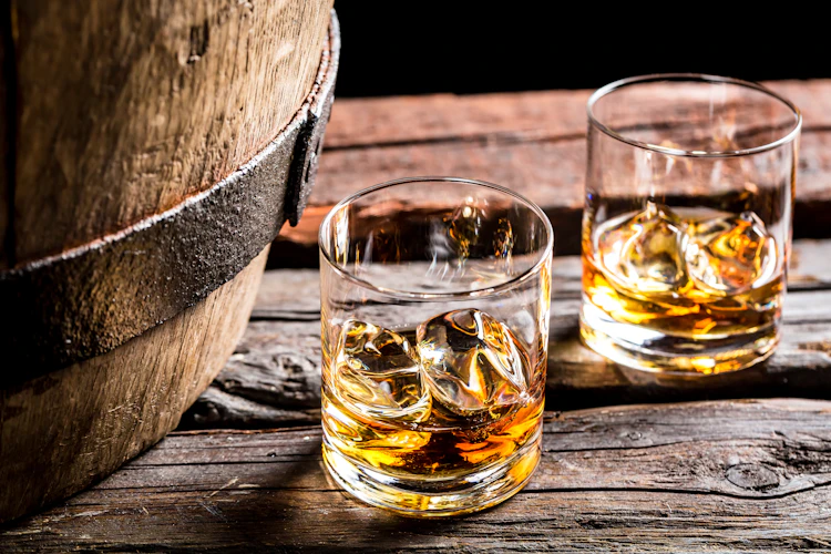 To glas med gylden whisky og isklumper på et rustikt træbord ved siden af et gammelt træfad i en traditionel kælder